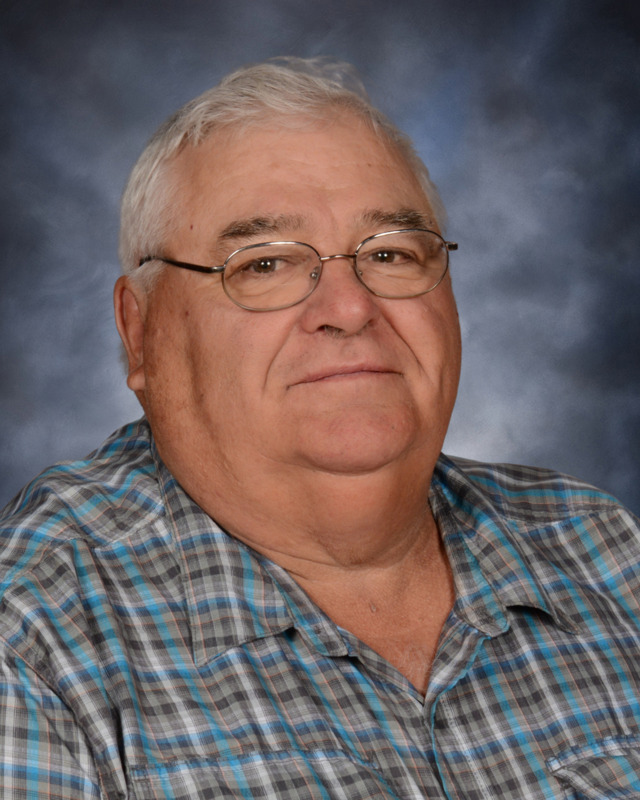 A middle-aged man with glasses and gray hair, wearing a plaid shirt, smiles at the camera against a softly blurred background.