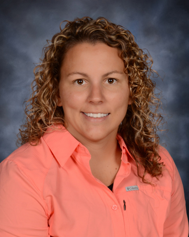A smiling woman with curly hair wearing an orange shirt, set against a blurred blue background.