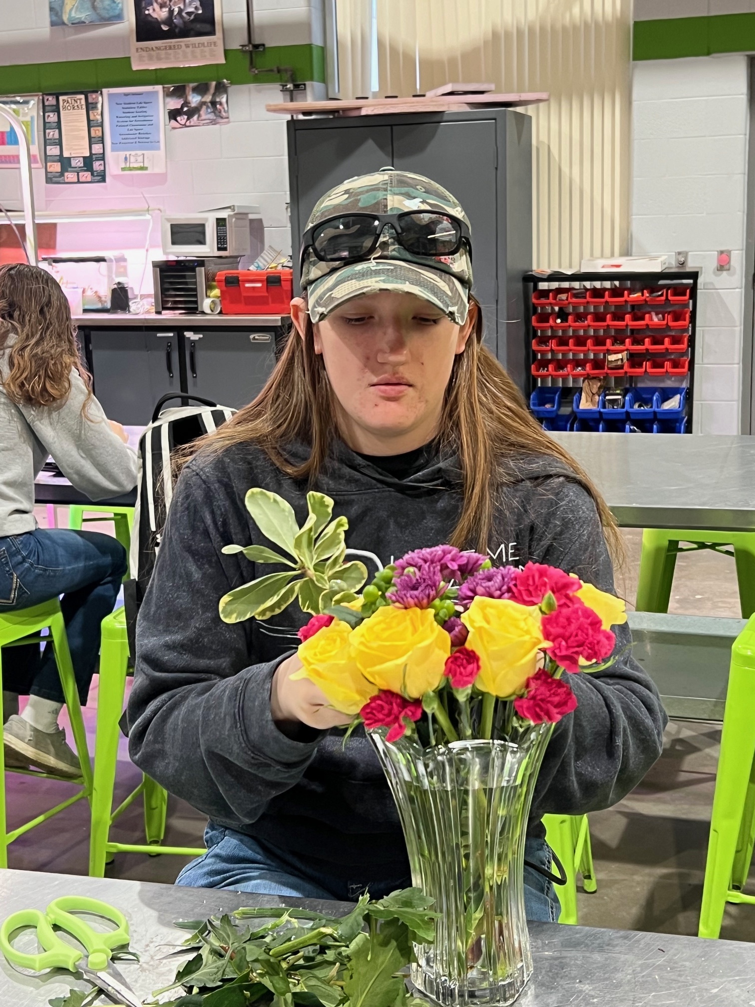 A person with long hair, wearing a cap and sunglasses, is arranging colorful flowers in a vase while seated at a table in a workshop setting.