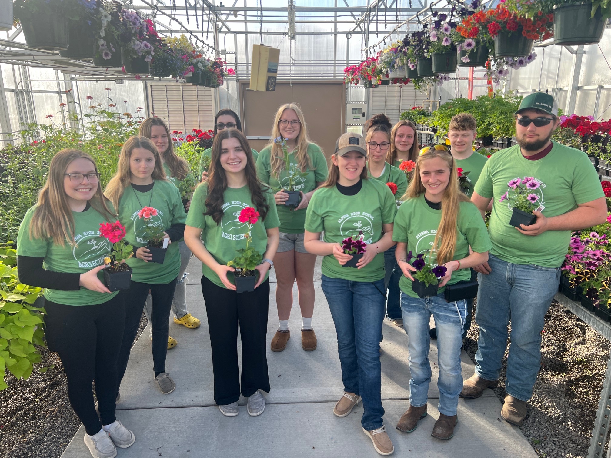 A group of twelve individuals wearing green shirts stands in a greenhouse, each holding a potted flower, surrounded by various plants and hanging flower baskets.