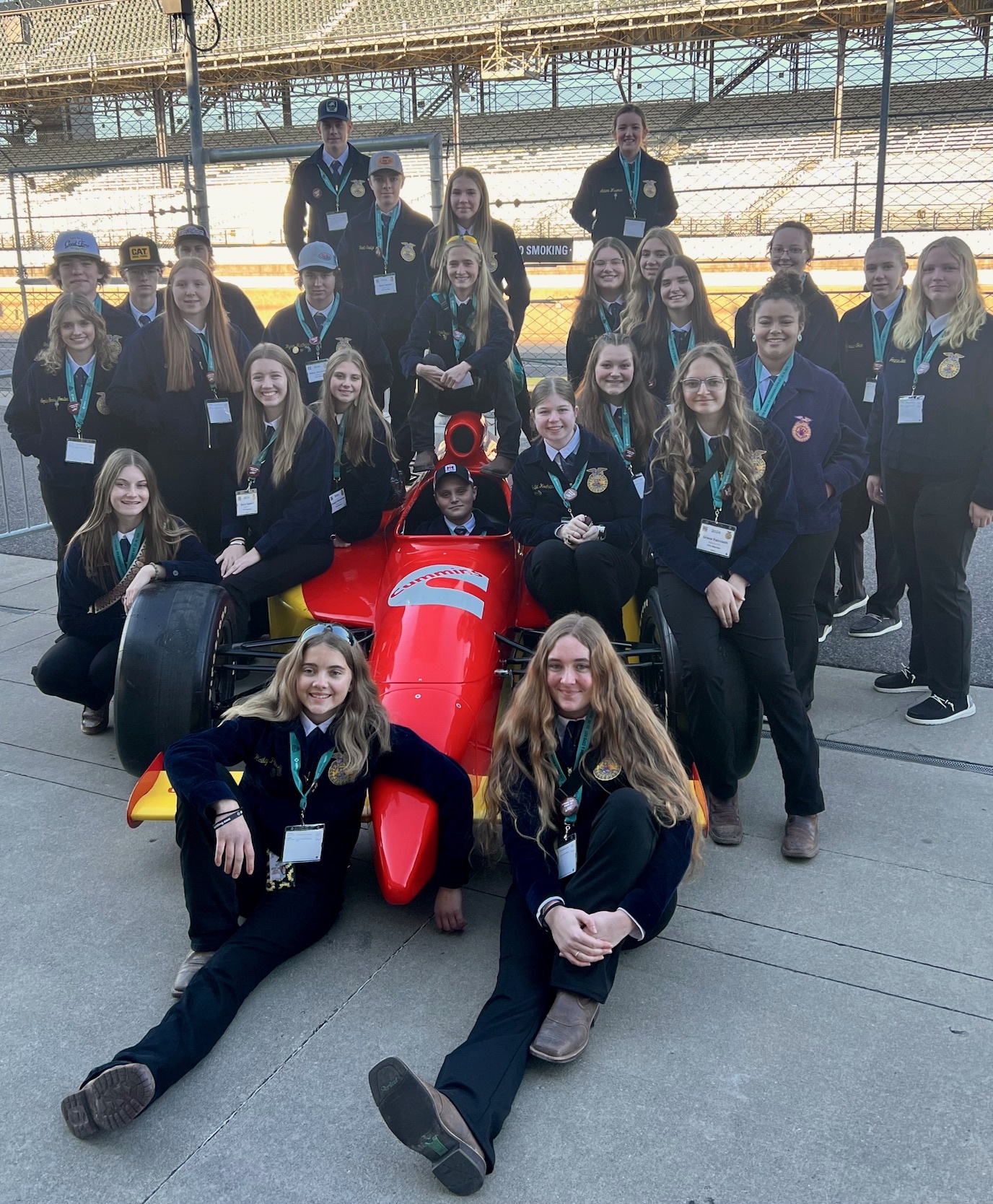 A group of young women in matching outfits poses together in front of a race car at a racetrack, with some standing and others sitting on the ground, all smiling and wearing identification badges.