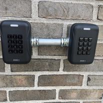 Two black keypad locks mounted on a brick wall, connected by a metal rod.