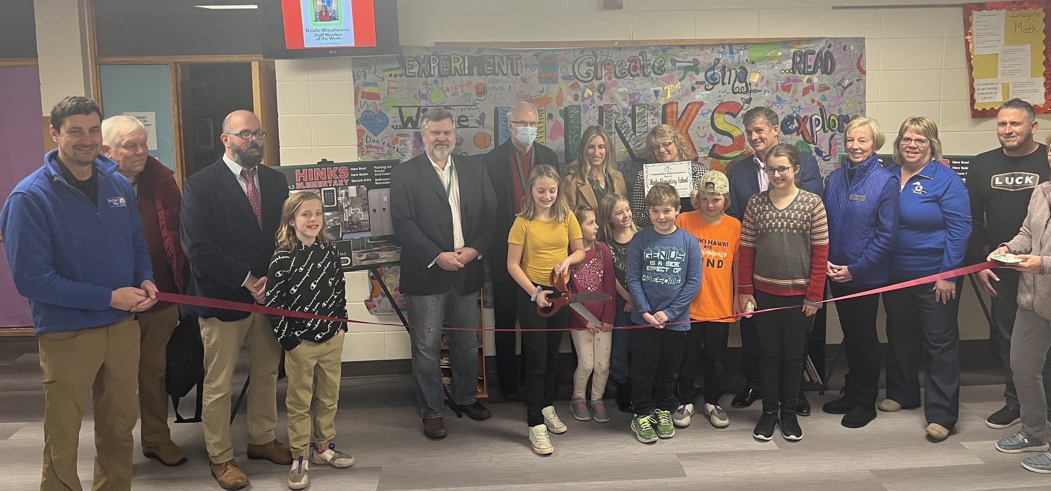 A group of adults and children gather for a ribbon-cutting ceremony, smiling and holding a large red ribbon in front of a colorful mural and display boards.