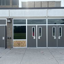 A view of a building entrance featuring multiple gray doors, with one door boarded up and a grassy area visible through the glass panels.