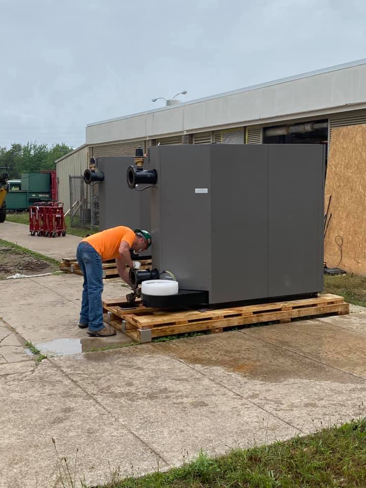 A worker in an orange shirt is crouched down, inspecting or working on a large gray equipment unit placed on a wooden pallet outside a building, with additional equipment visible in the background.