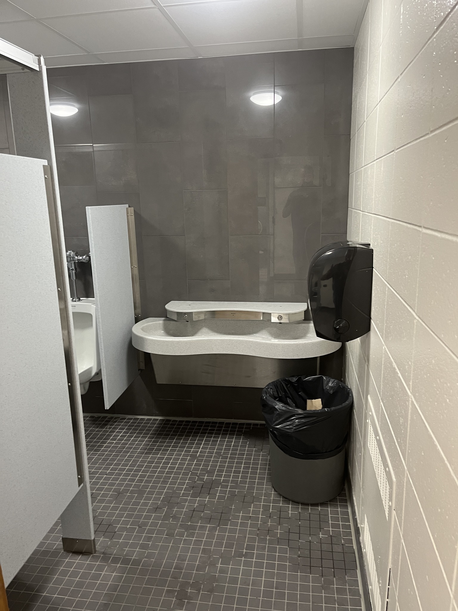 A modern restroom interior featuring a sink, soap dispenser, and trash can, with gray tiled walls and floor.