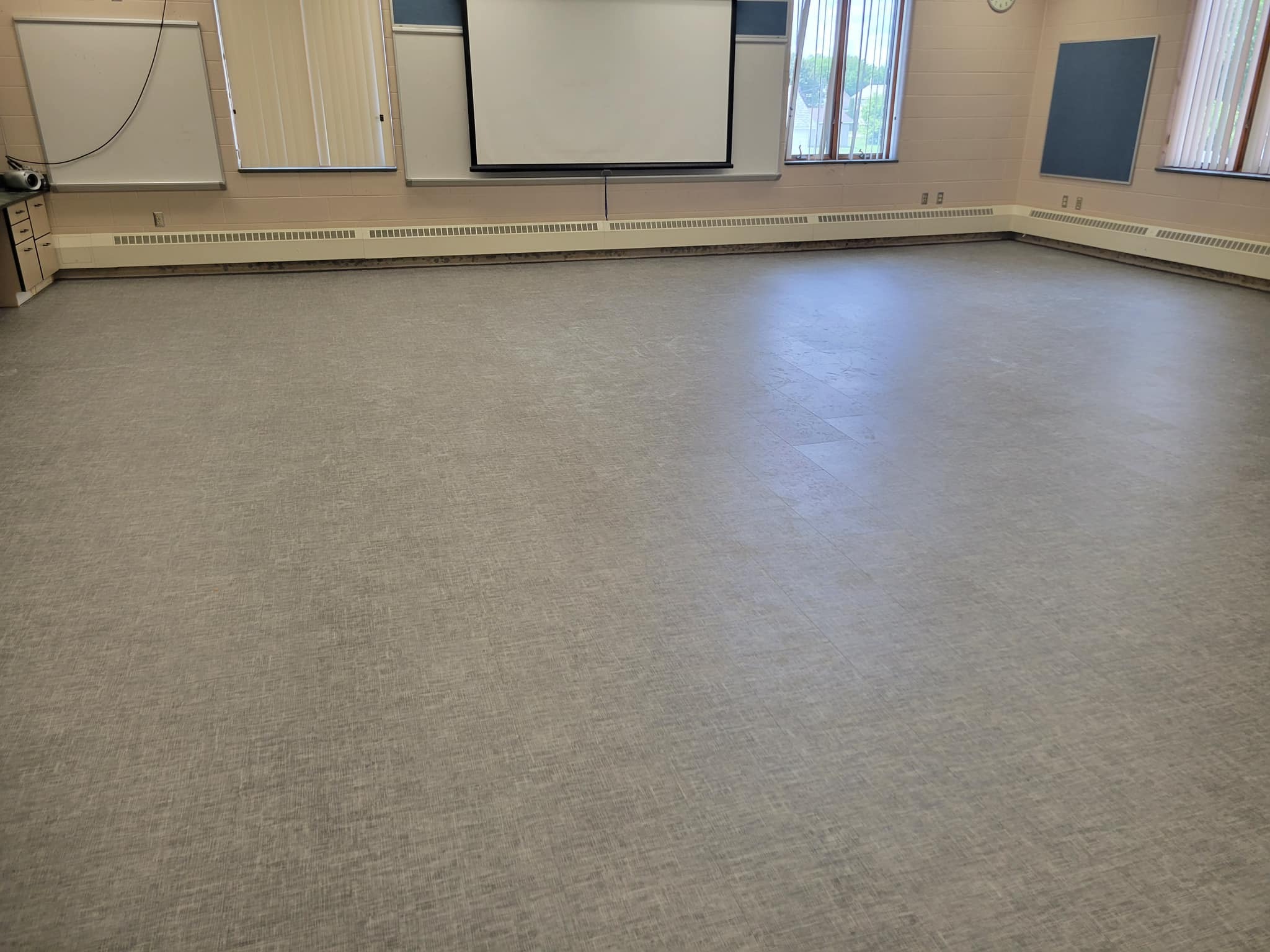A spacious, empty room with a gray floor, large windows allowing natural light, and a projection screen on the front wall.