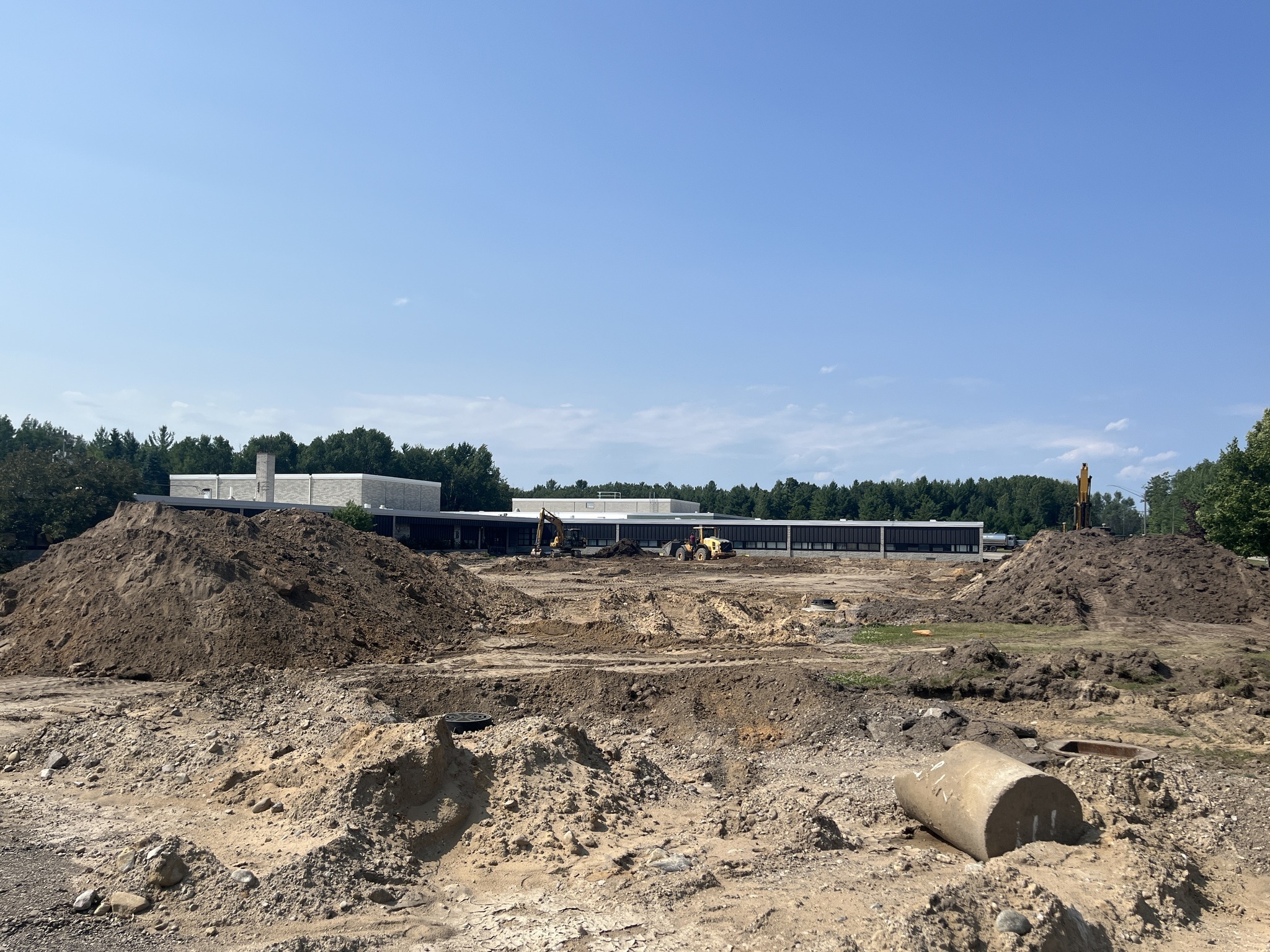 A construction site featuring large mounds of dirt and machinery, with a partially built structure in the background under a clear blue sky.