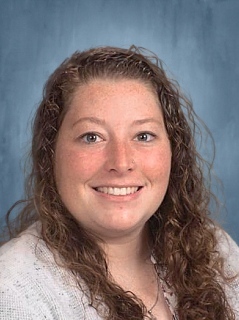 A smiling woman with curly hair, wearing a light sweater, poses for a portrait against a blue background.
