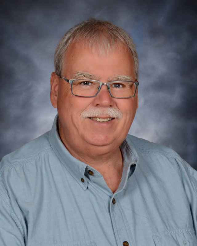 A smiling older man with glasses and a mustache, wearing a light blue shirt, against a softly blurred gray background.