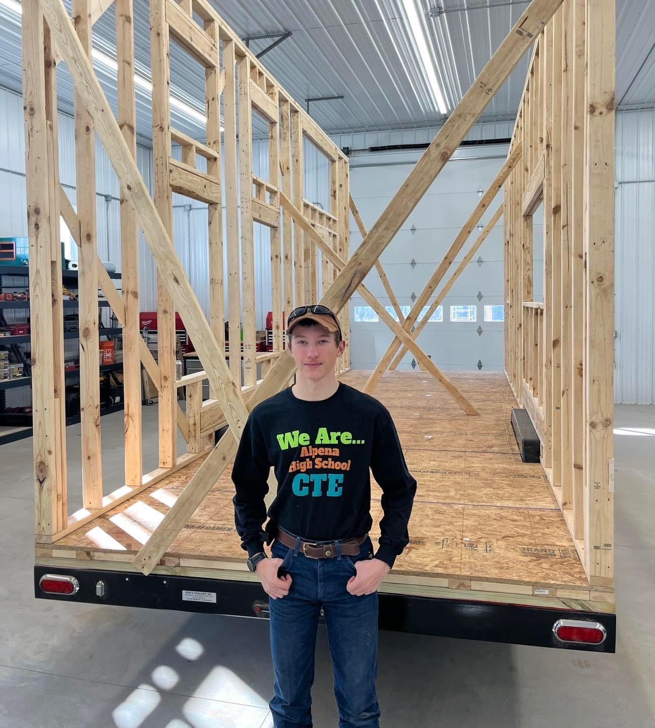 A young person stands in front of a partially constructed wooden frame on a trailer, inside a spacious building with bright lighting.