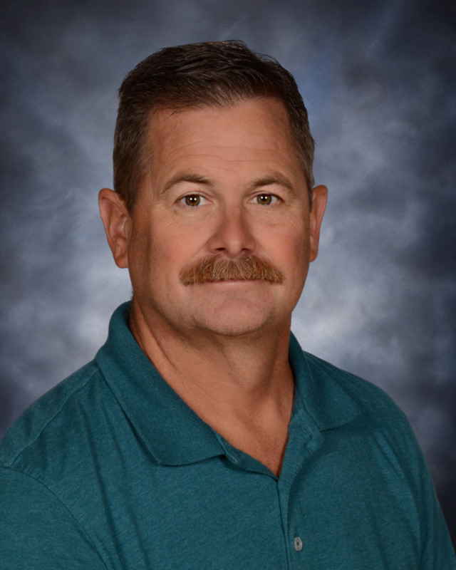A man with a mustache is wearing a teal polo shirt, posing against a softly blurred gray background.