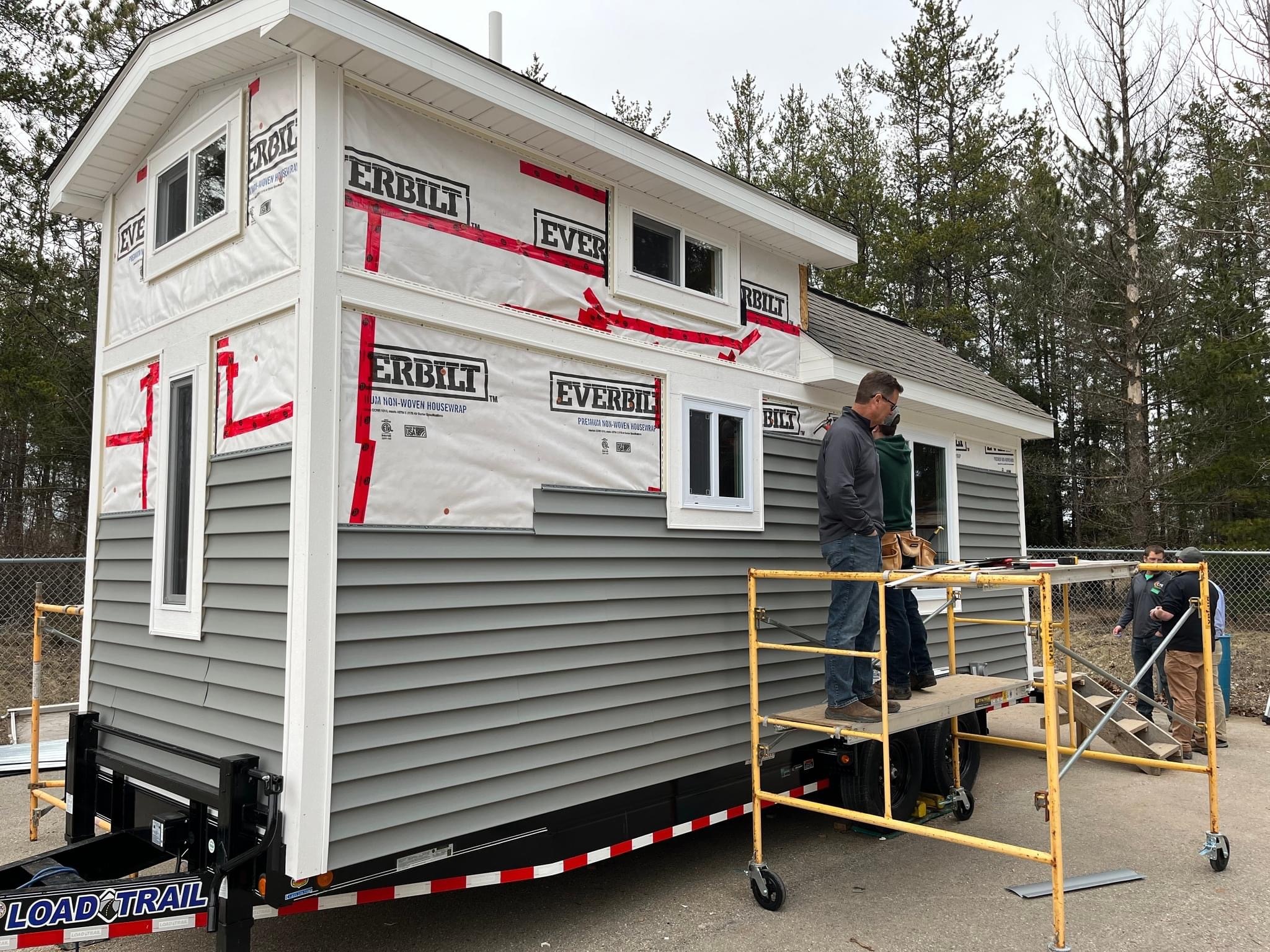 A man is working on the exterior of a tiny house that is partially wrapped in protective material, with scaffolding set up beside it, surrounded by trees.
