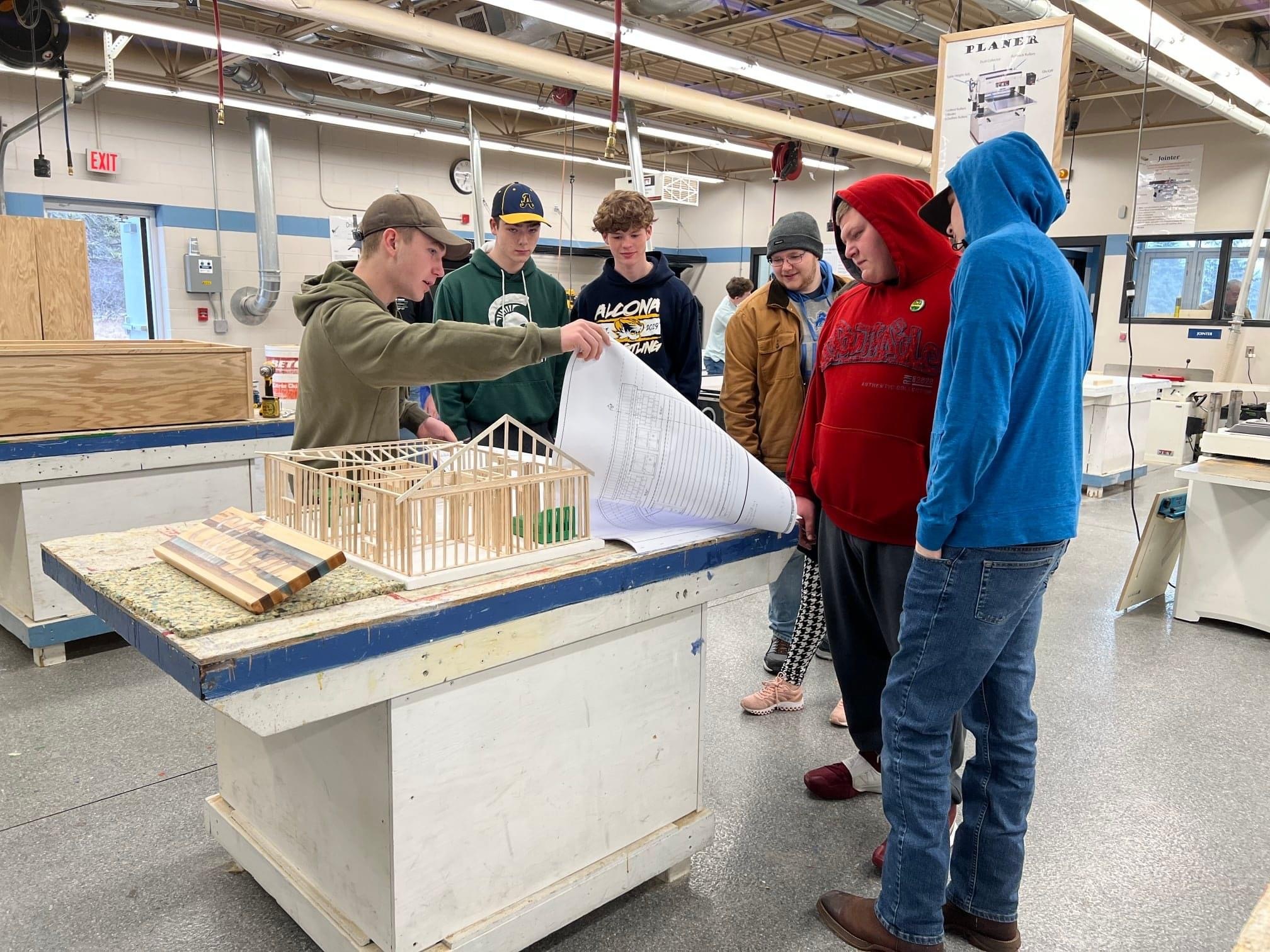 A group of students gathers around a table in a workshop, observing a teacher as he demonstrates a construction project involving a model house and blueprints.