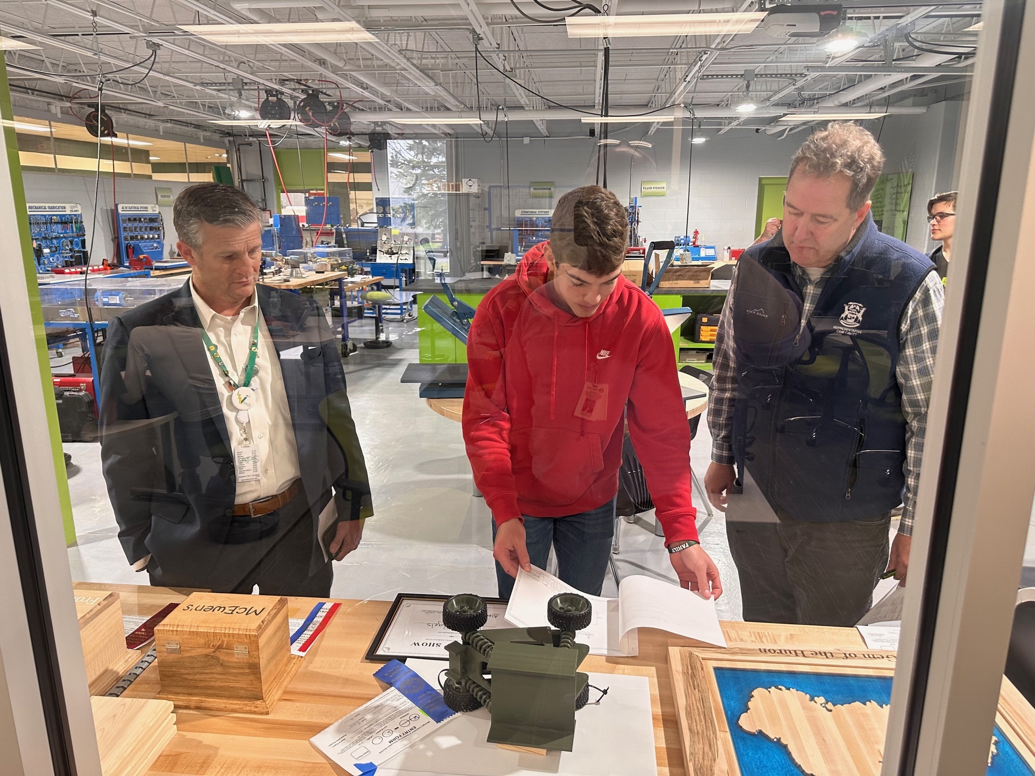 Three individuals are observing and discussing materials on a table in a workshop or lab setting, with various tools and equipment visible in the background.