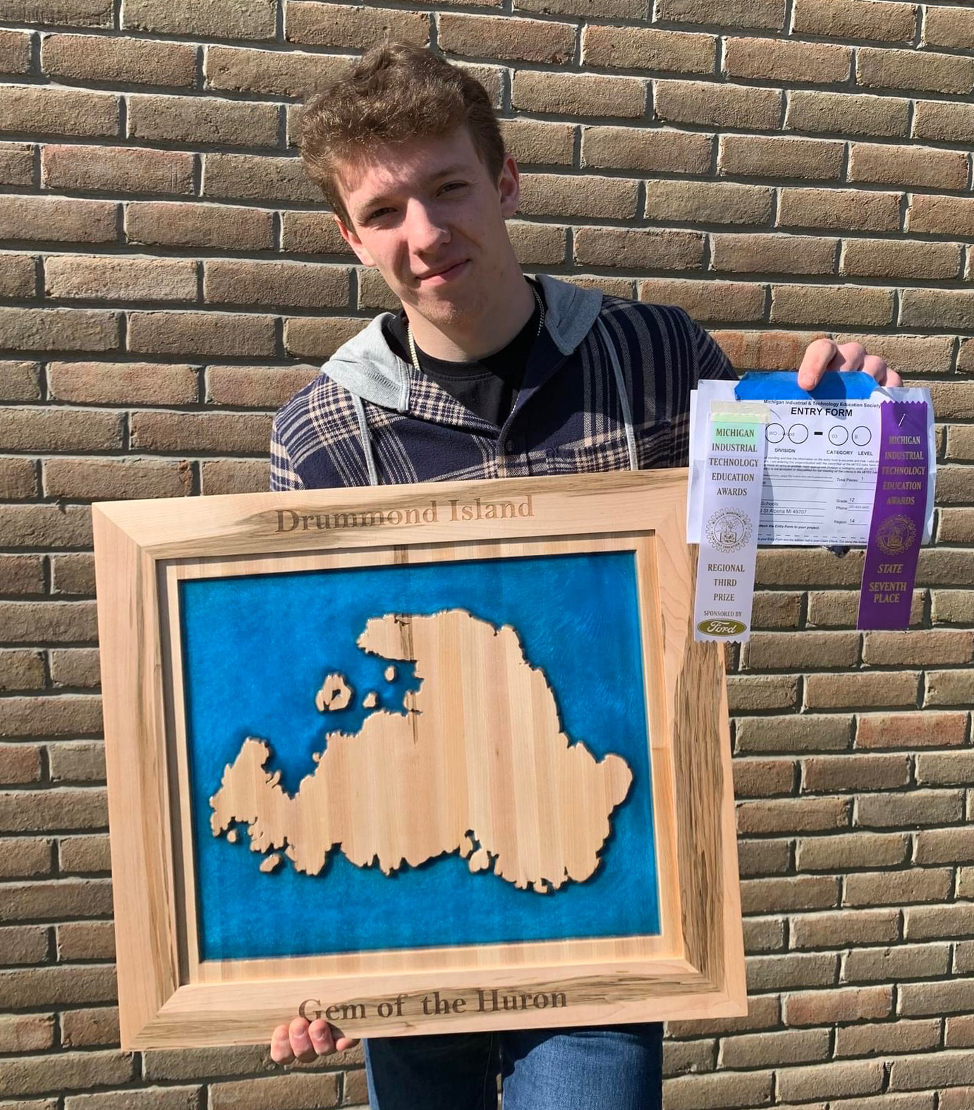 A young man holds a framed wooden map of Antarctica, showcasing its shape against a blue background, along with several ribbons and certificates.