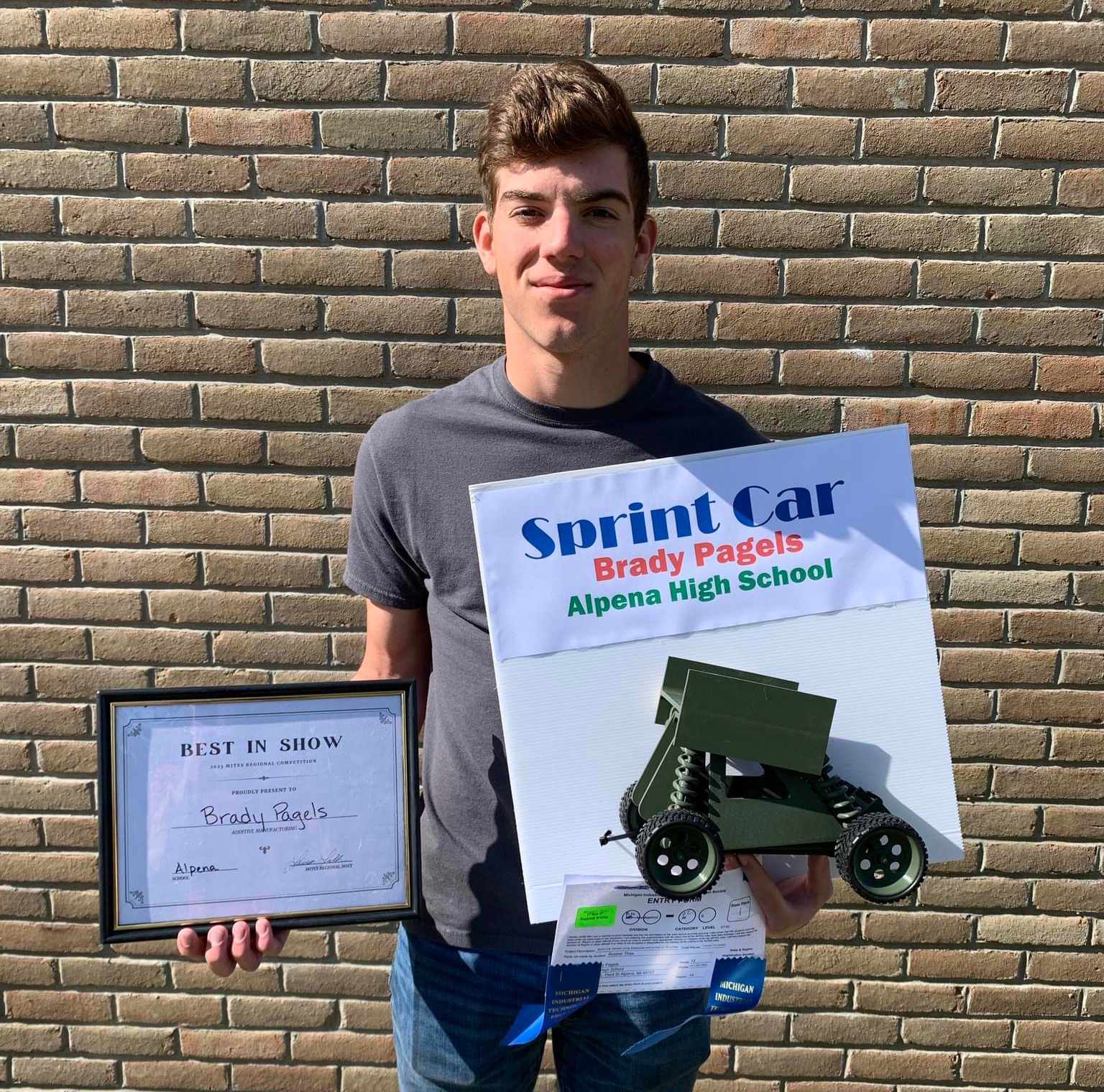 A young man stands in front of a brick wall, holding a certificate and a large sign that reads 'Sprint Car' along with a model of a sprint car, representing an achievement from Algena High School.