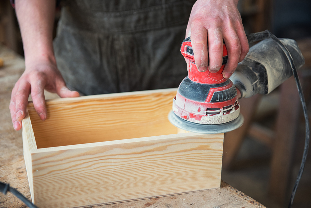 A person is using a power sander on a wooden box, smoothing its surface in a workshop setting.