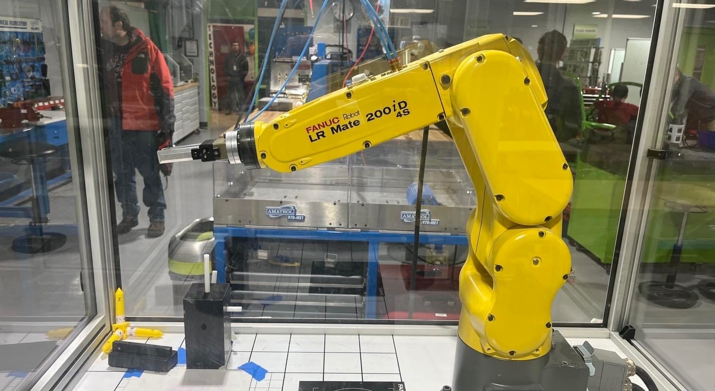 A yellow robotic arm is positioned inside a glass enclosure, with a person visible in the background observing the setup.