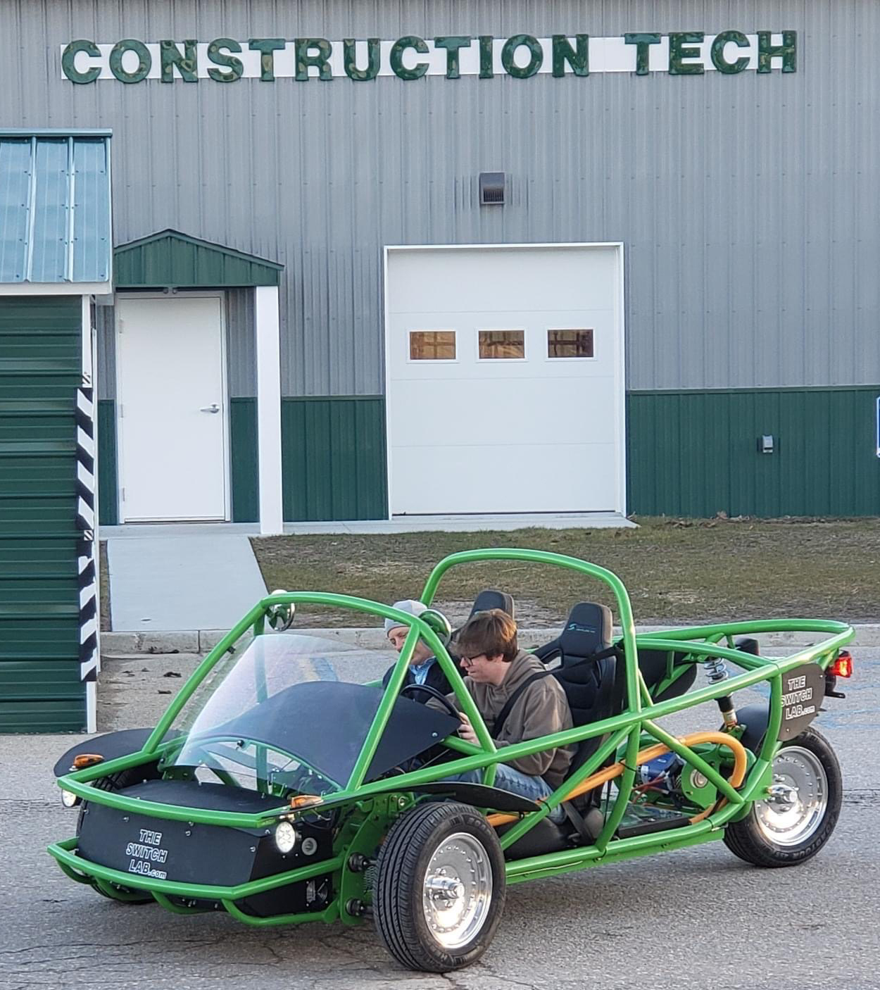 A person is seated in a green and black open-frame vehicle outside a building with a sign that reads 'CONSTRUCTION TECH'.