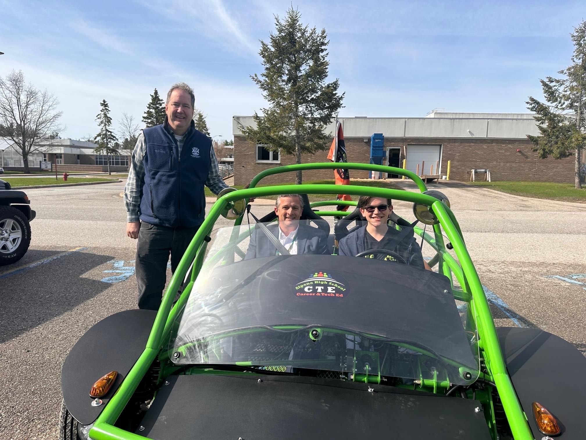Three people are posing in and around a green off-road vehicle, with a clear sky and a building in the background.