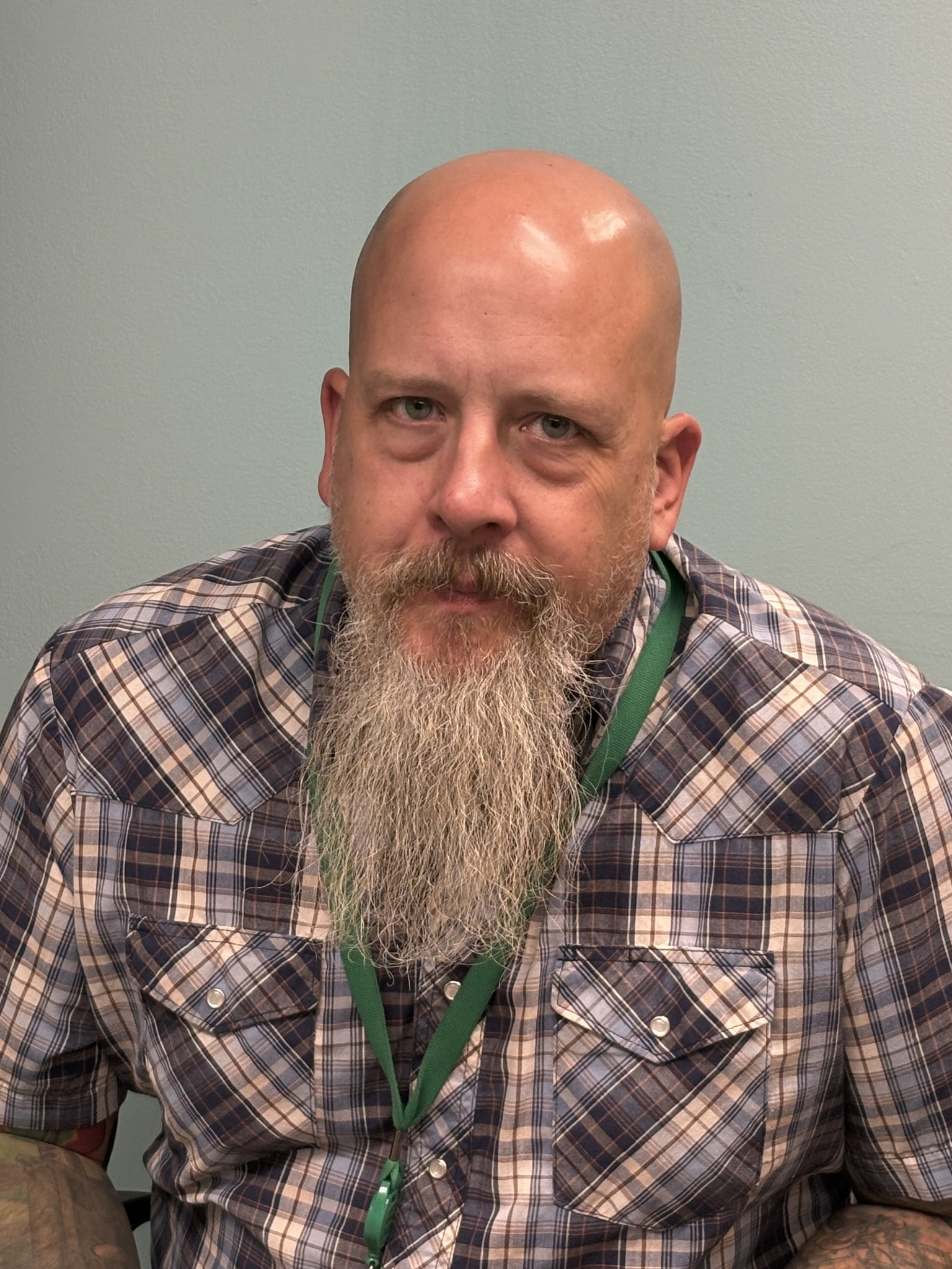 A man with a bald head and a long beard, wearing a plaid shirt and a green lanyard, is sitting against a light blue background.