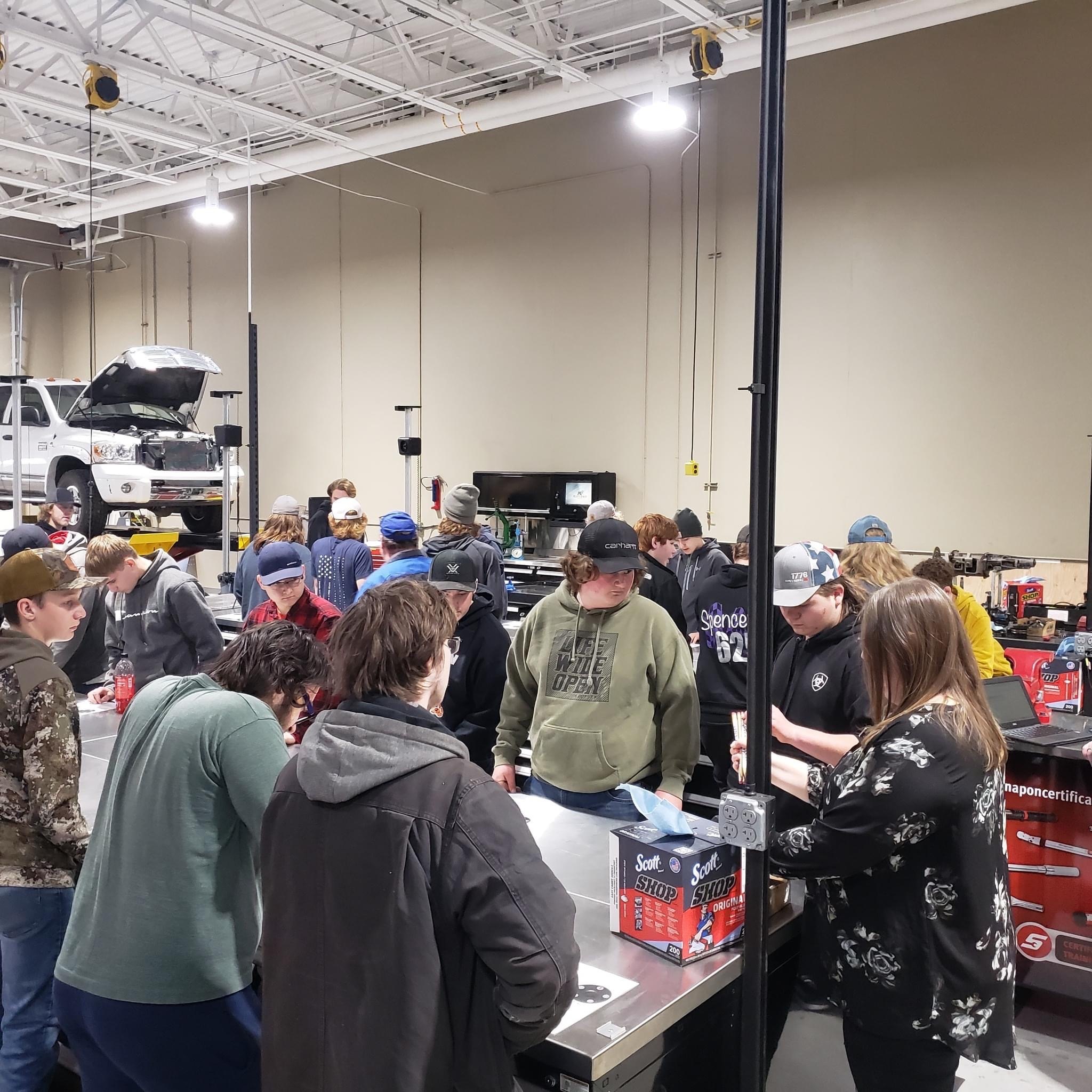 A group of people gathered around tables in a workshop, engaged in discussions and activities related to automotive tools and equipment, with vehicles visible in the background.