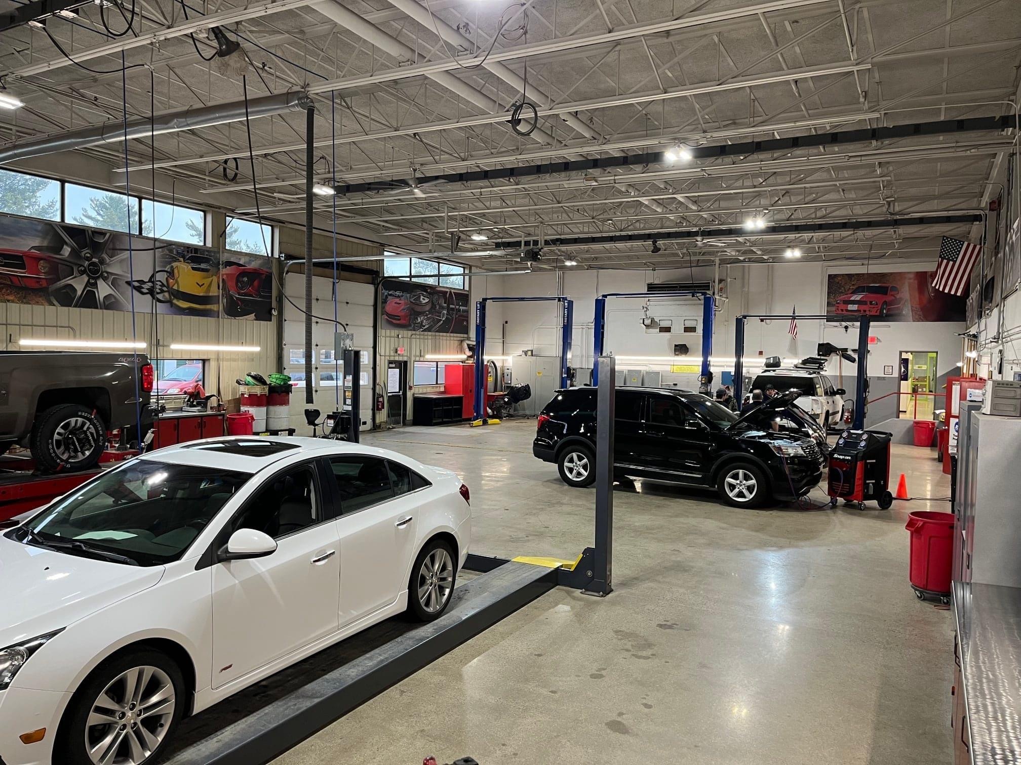 A spacious automotive repair shop featuring multiple service bays, with a white car on a lift in the foreground and a black SUV being serviced in the background.