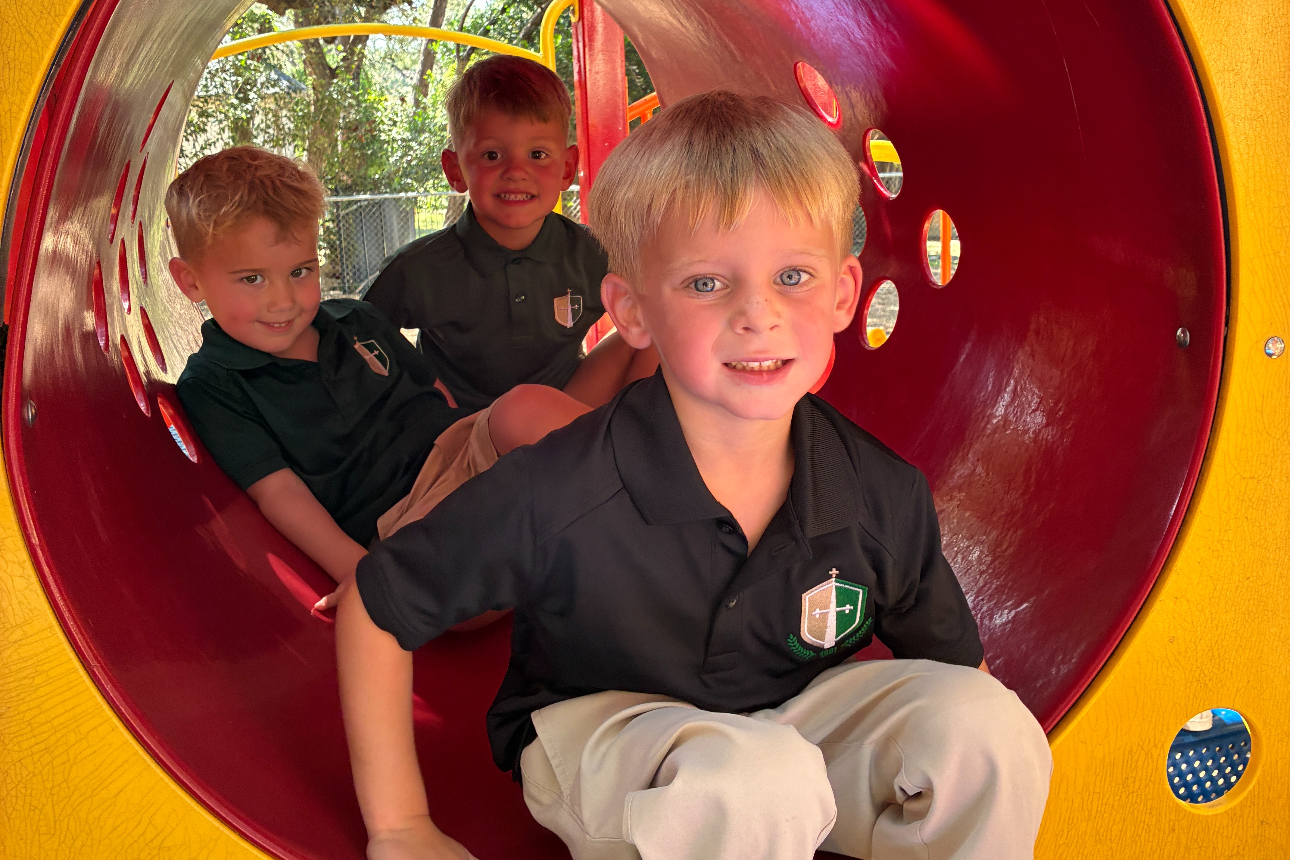 Three young boys are sitting inside a colorful, circular tunnel on a playground. Trees are visible in the background.