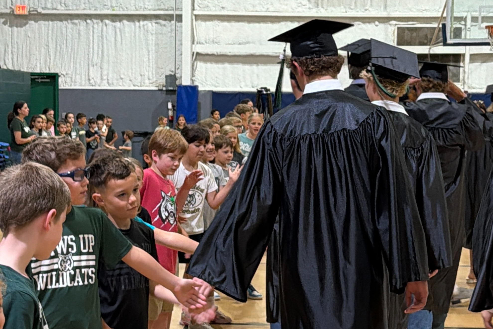 Several young children and a few adults in gowns and hats walking in a line across a basketball court.