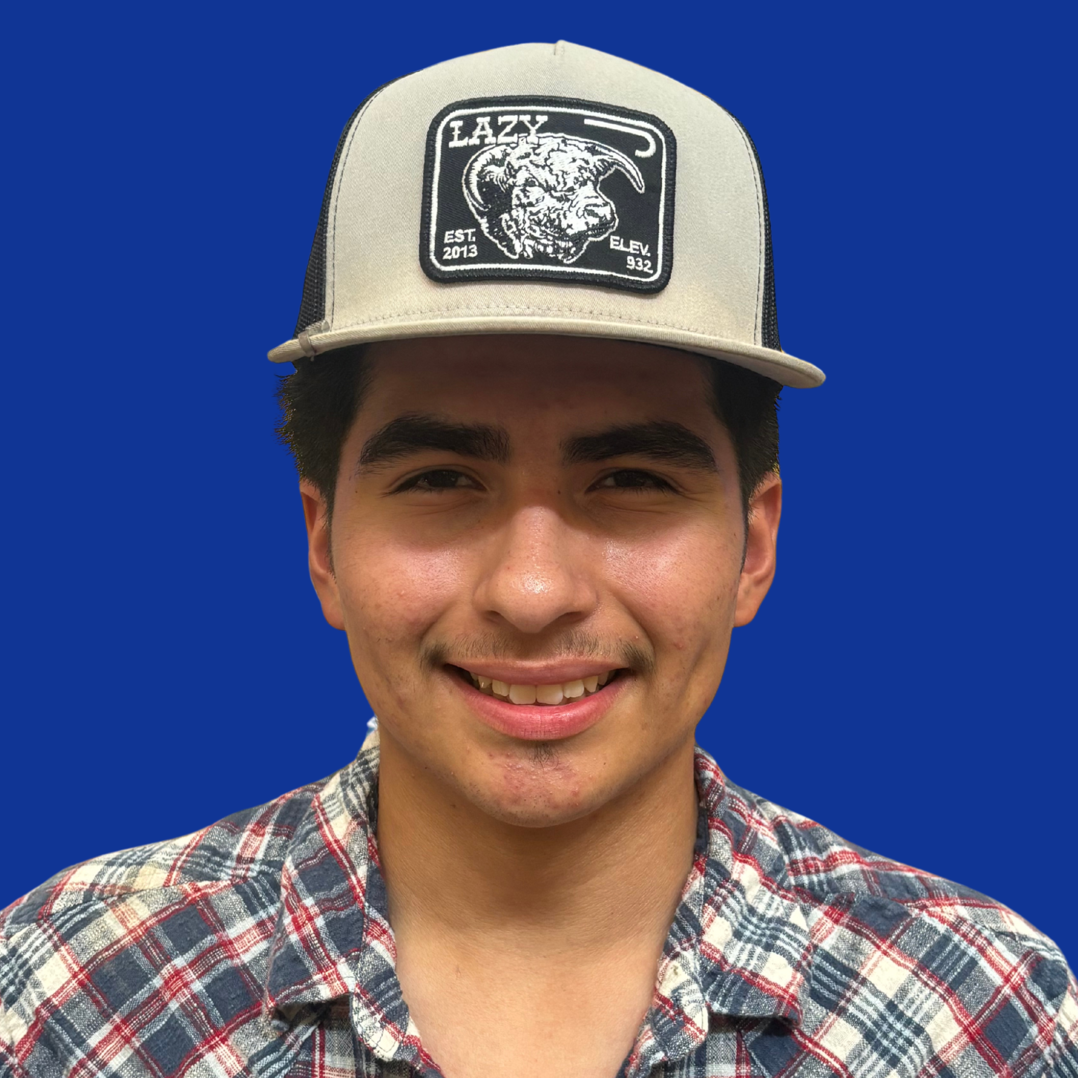 A person stands against a blue background, wearing a plaid shirt and a cap with a black design.