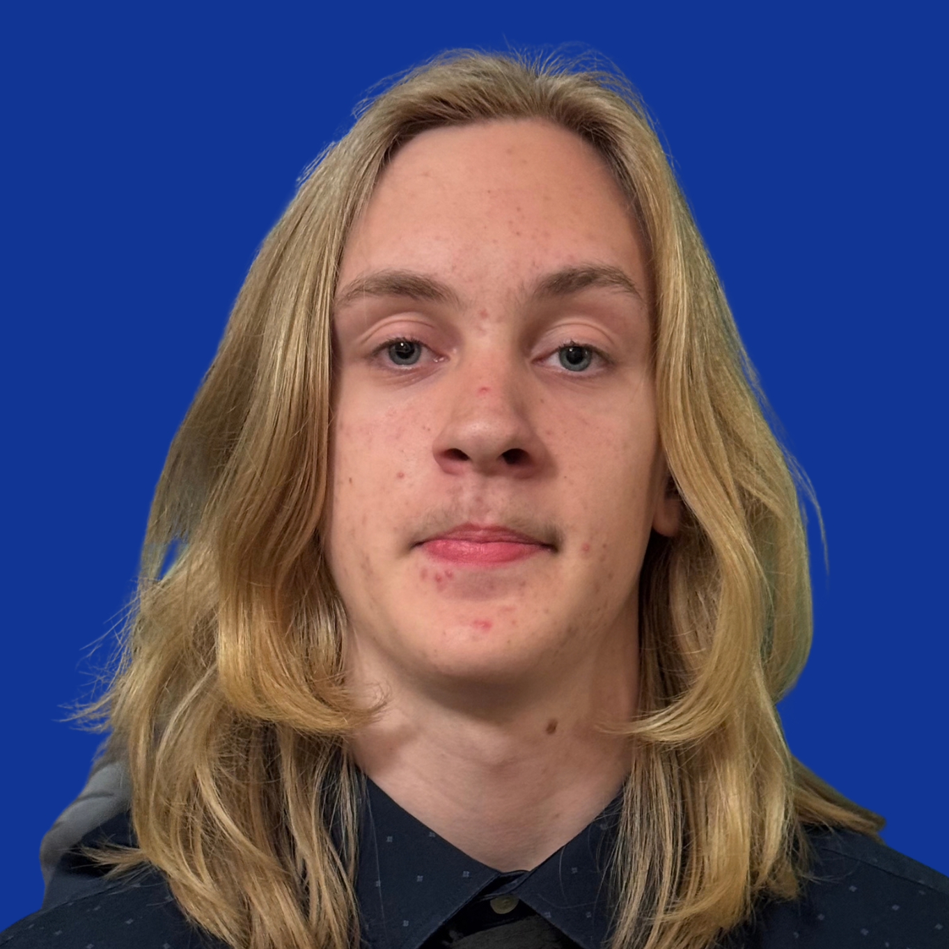 A person stands against a blue background, wearing a plaid shirt and a cap with a black design.