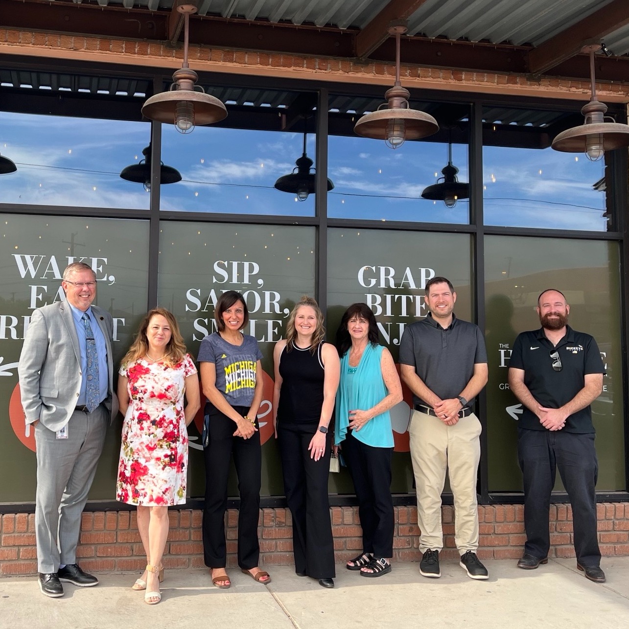 A group of people stands outside a brick building with large windows and glass doors. The sign on the door says "WAKE UP" and "SIP".