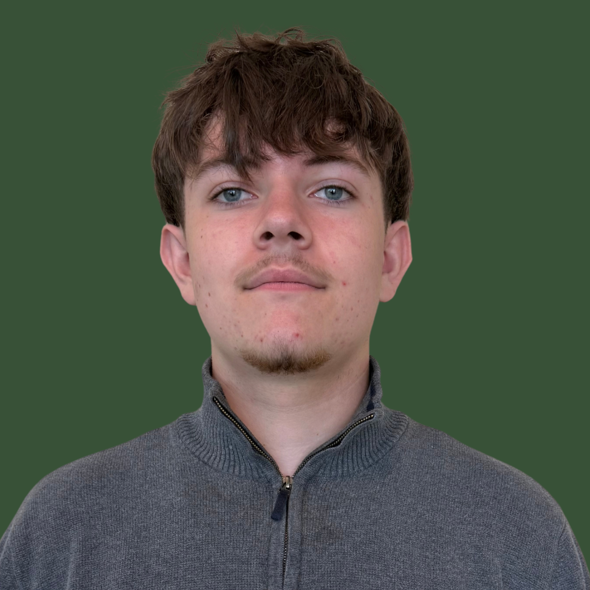 A man with short, dark hair and a light beard stands against a green background. He wears a gray sweater with black stripes.