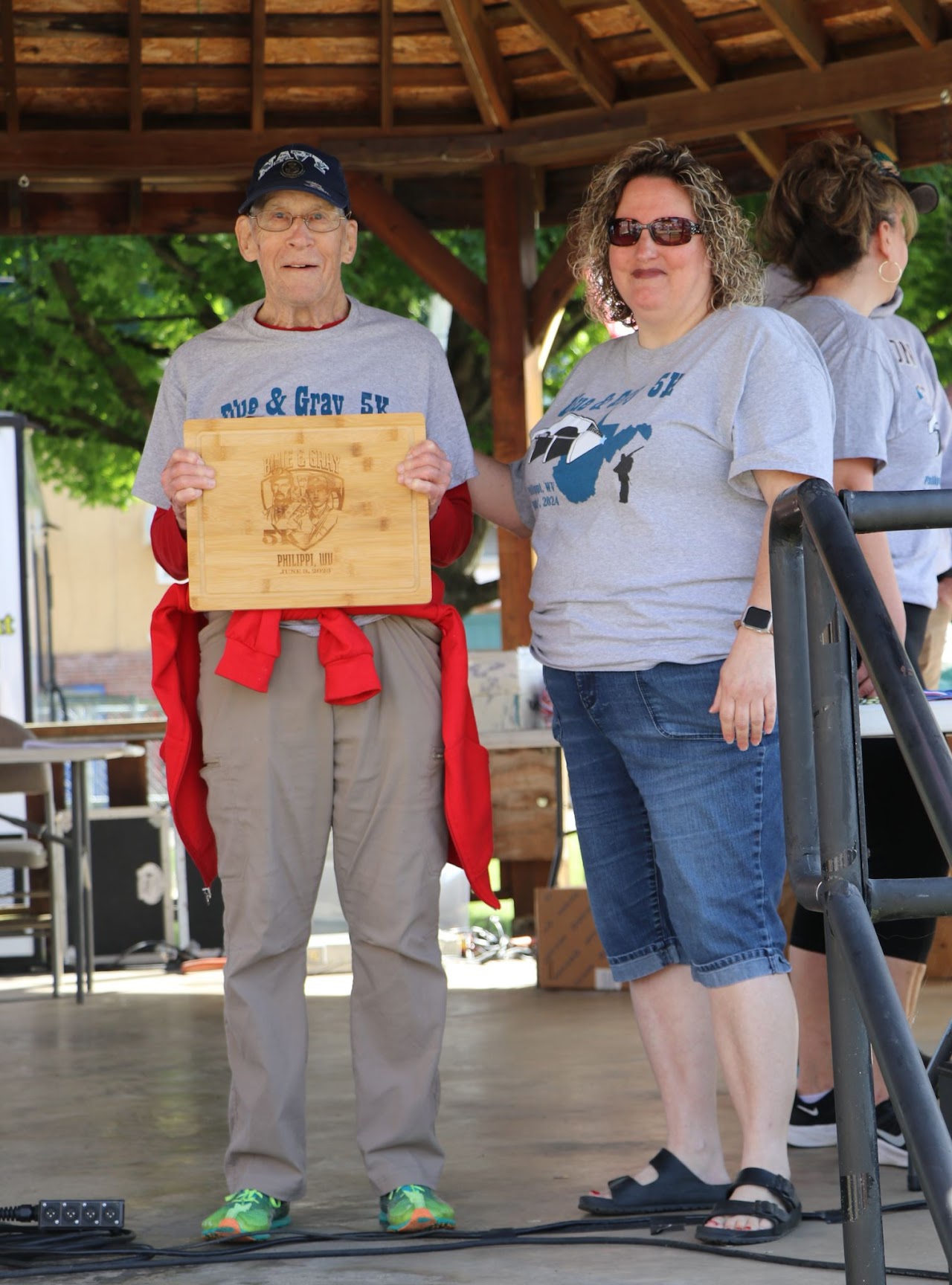 Two participants at an award ceremony, one holding a wooden plaque, standing beneath a wooden structure with green trees in the background.