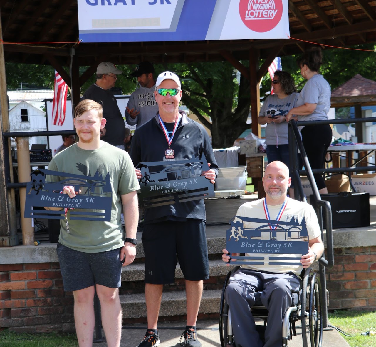 Three participants proudly display their awards at the Blue & Gray 5K event in Philippi, WV, under a decorated pavilion.