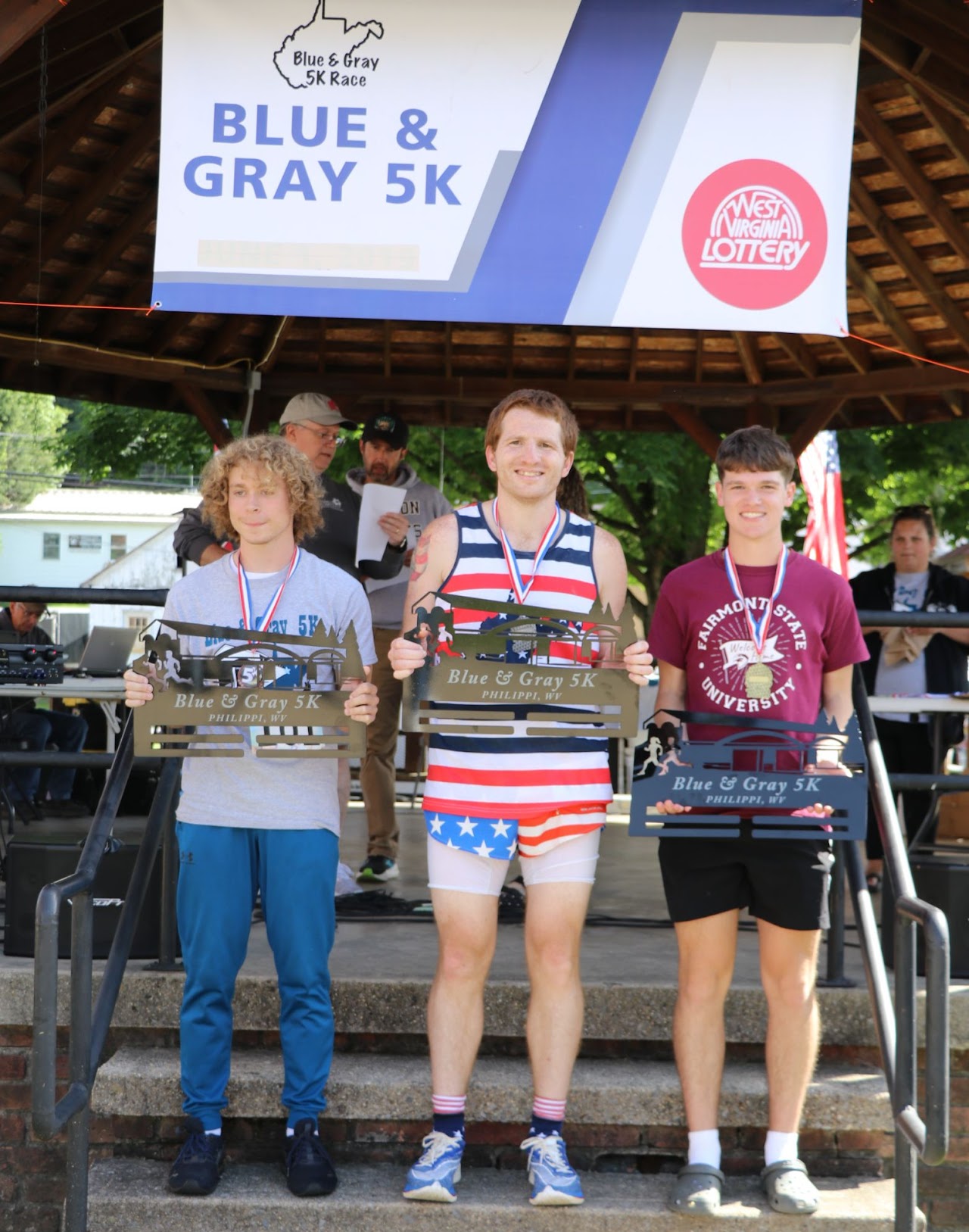 Three male runners proudly display their awards after the Blue & Gray 5K race, adorned with medals and standing on a stage.