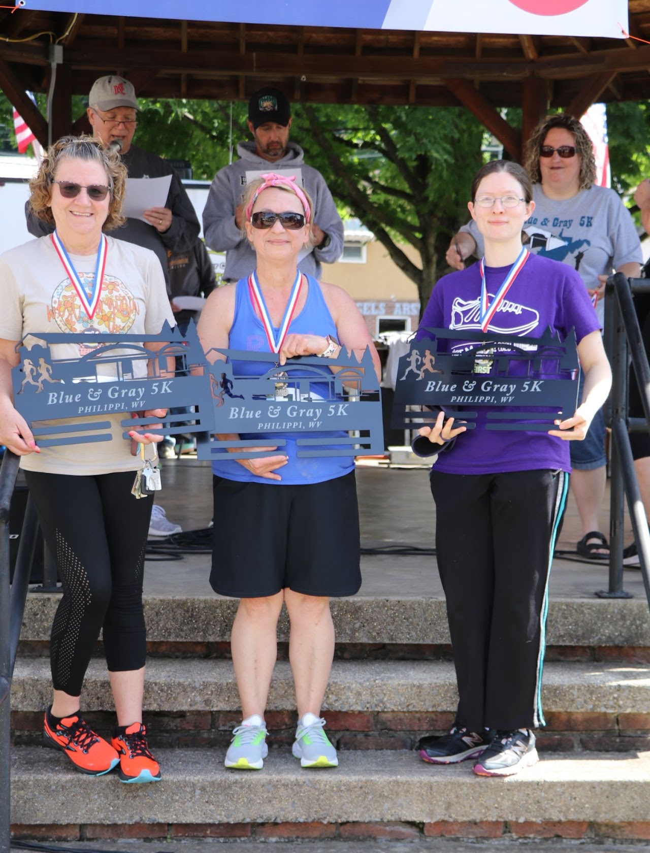 Three participants proudly display their awards for the Blue & Gray 5K, wearing medals and athletic gear, on a stage outdoors.