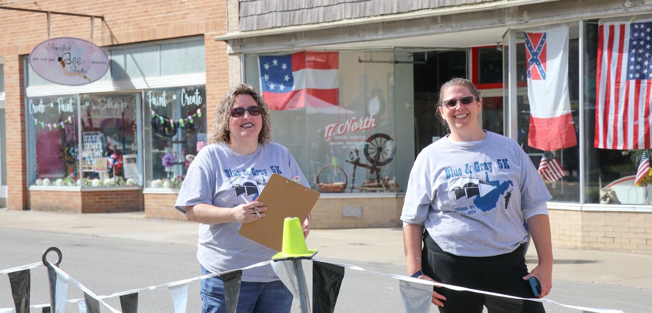 Two women in "Blue & Gray 5K" shirts stand outside a shop, one holding a clipboard and a yellow cone, with flags in the background.