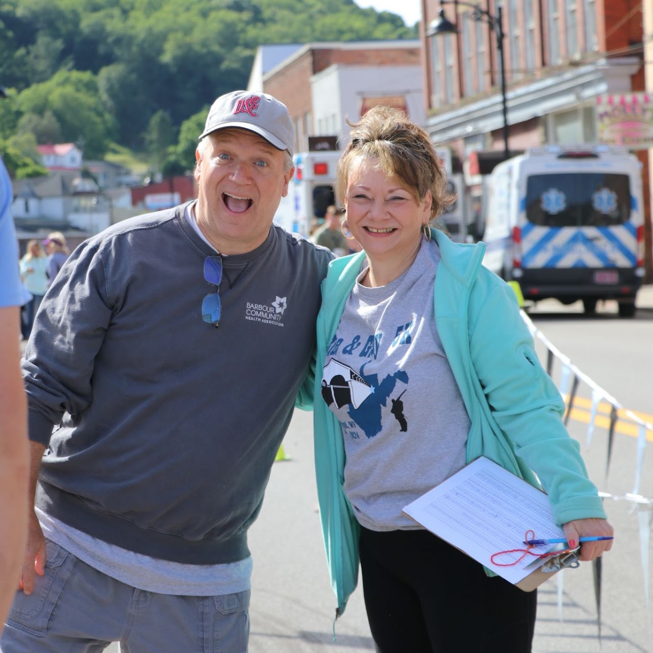 Two people pose together outdoors, smiling, with a clipboard in one person's hand. Colorful storefronts and trees are in the background.