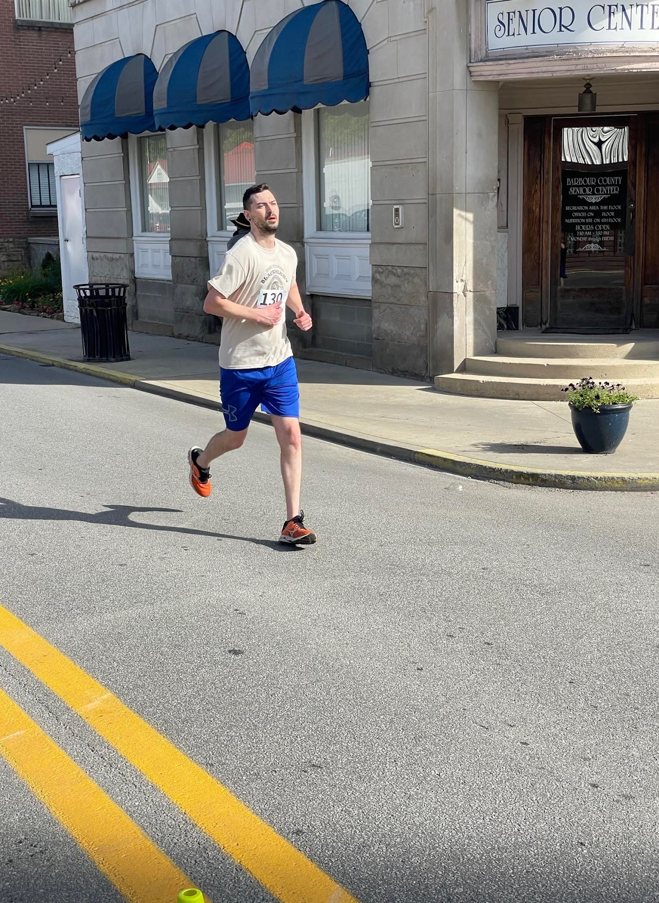 A runner wearing a beige t-shirt and blue shorts strides down a street lined with a senior center and shops.