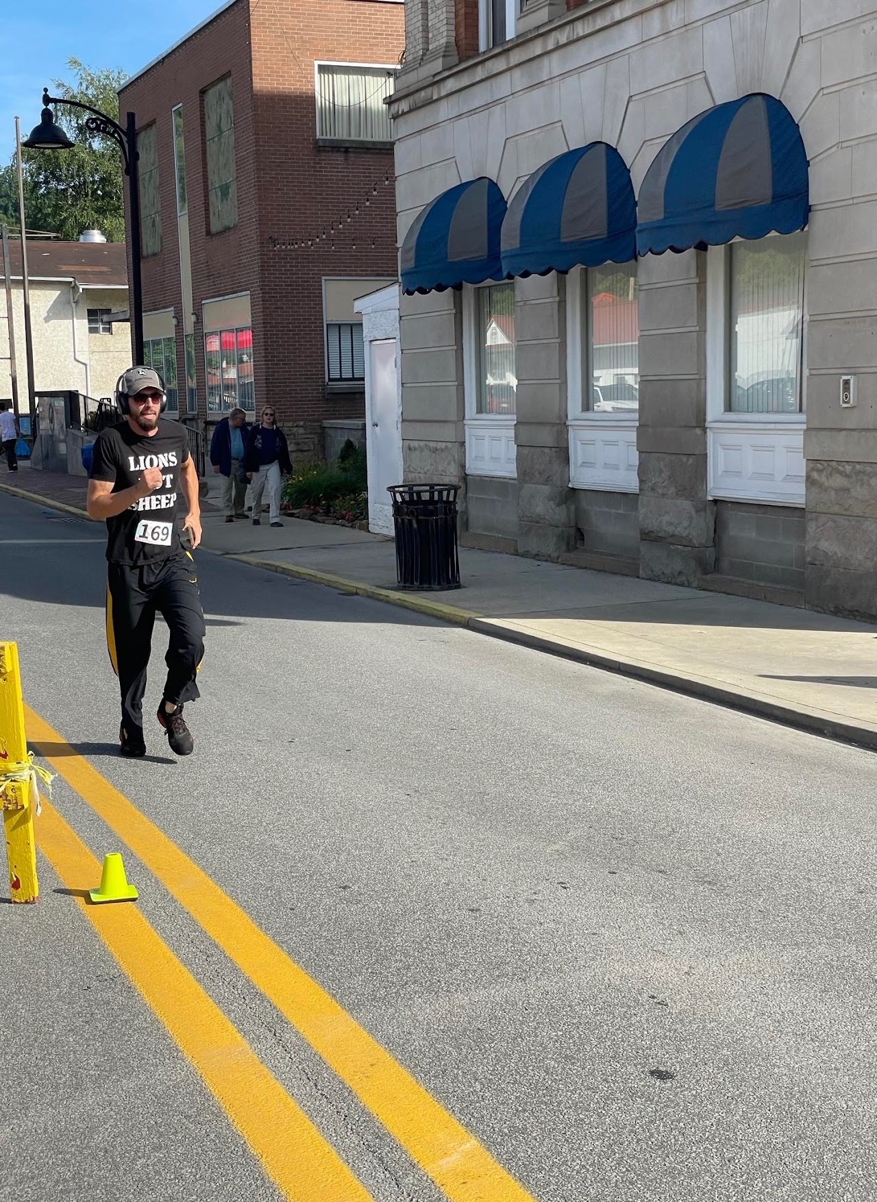 A runner in a black "LIONS NOT SHEEP" shirt, numbered 169, jogs down a street lined with buildings and spectators.