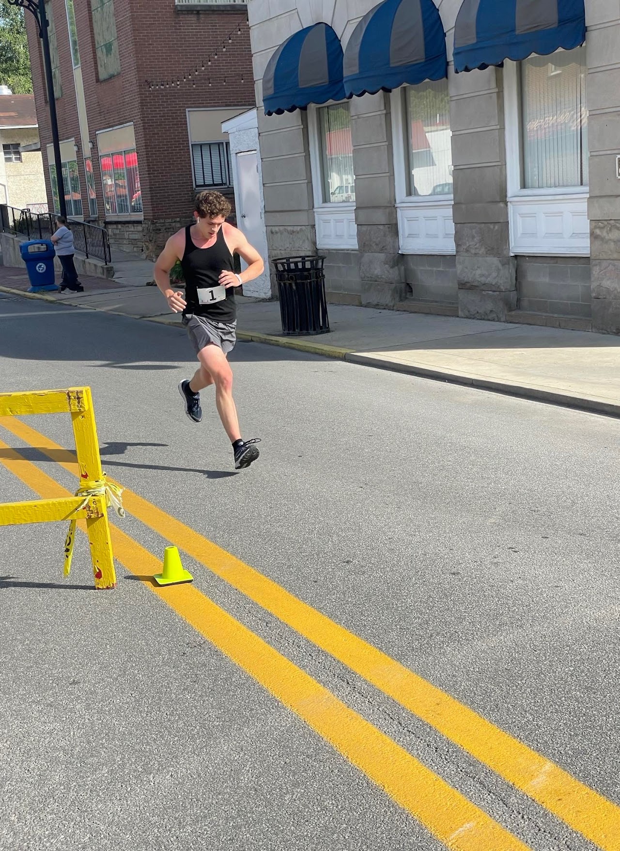 A runner in shorts and a tank top sprints down a street, navigating a yellow barrier with a cone beside him. Buildings line the background.