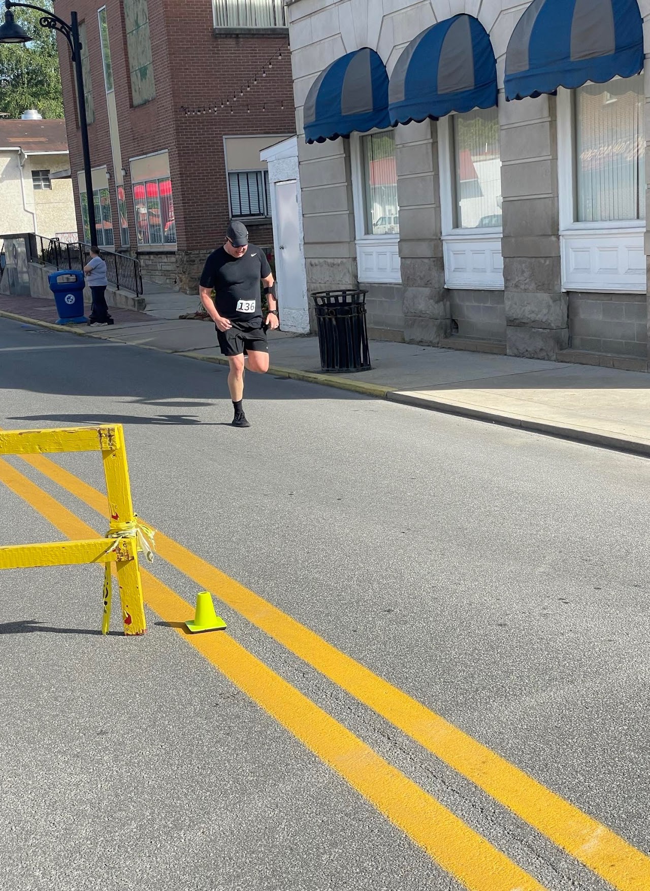 A runner in black athletic gear races down a city street, passing a yellow barrier and a green traffic cone, with buildings in the background.