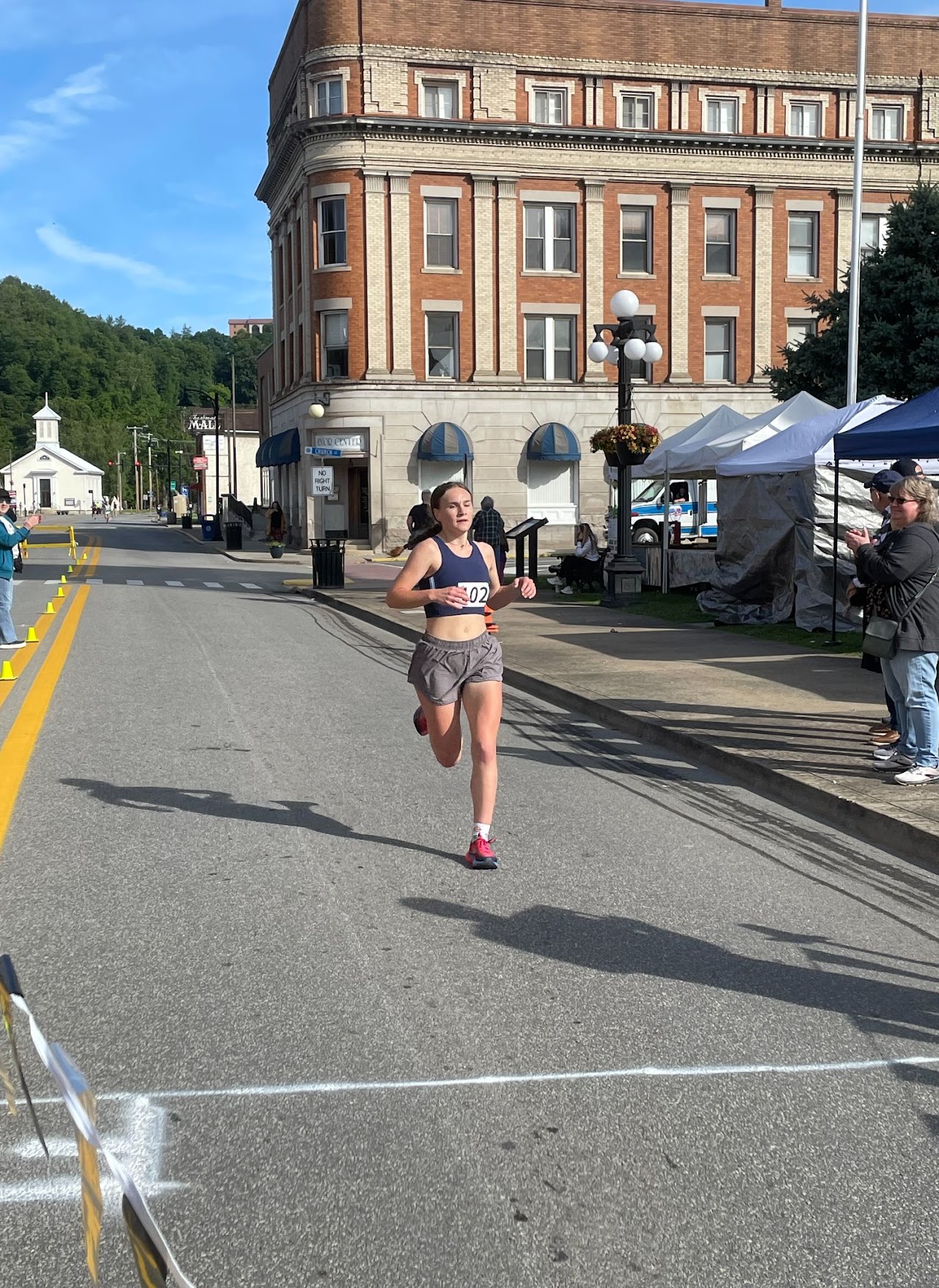 A runner in athletic gear dashes down a street during a race, with spectators and a historic building in the background.