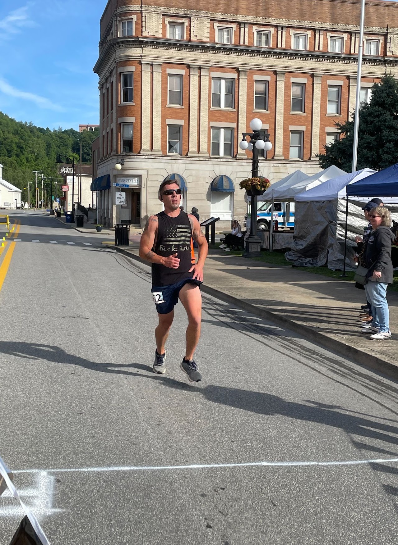 A runner in athletic gear crosses the finish line on a city street, with spectators and vendor tents in the background.