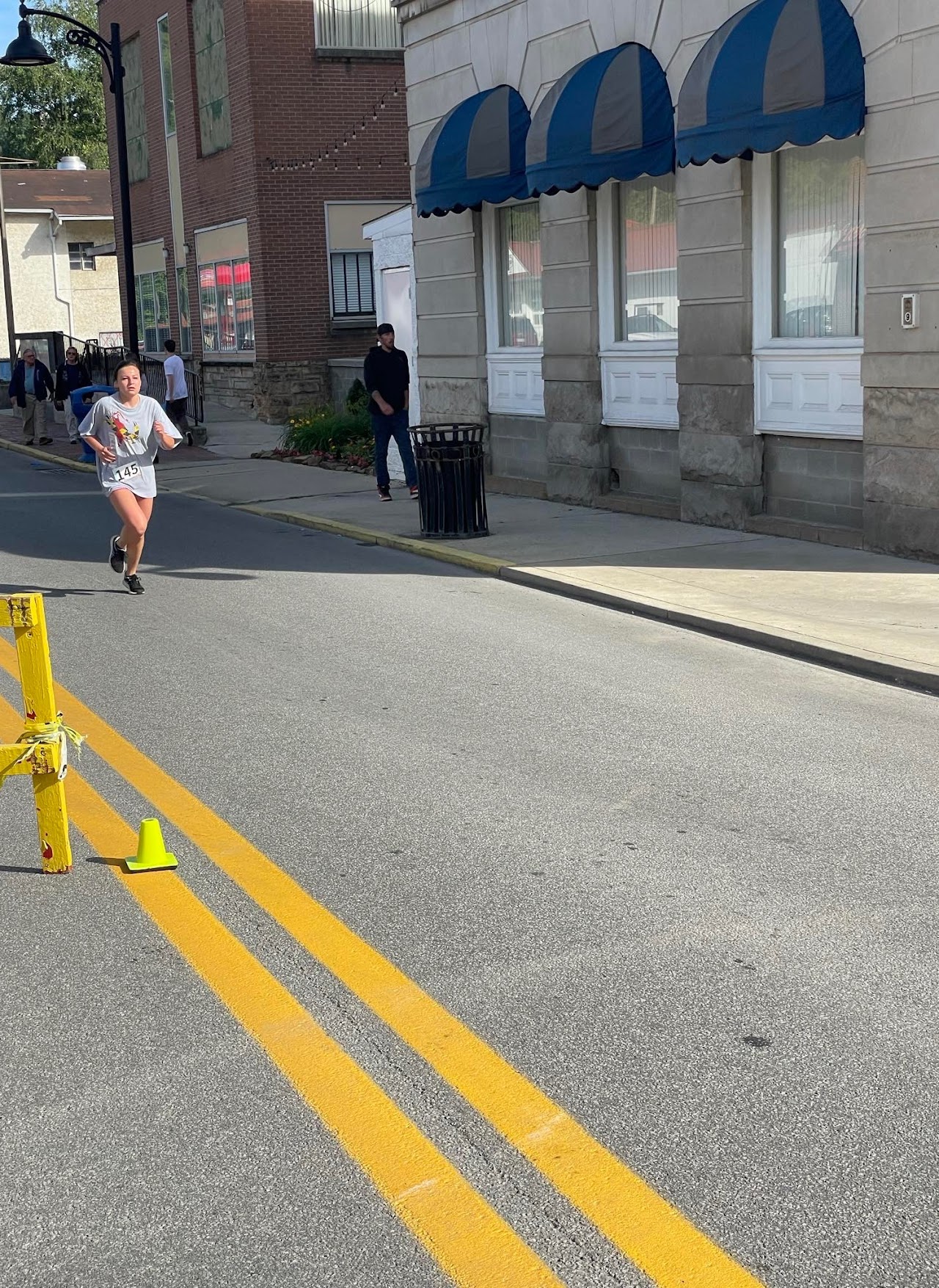 A runner in a numbered shirt sprints past a yellow barricade on a city street, with bystanders and buildings in the background.