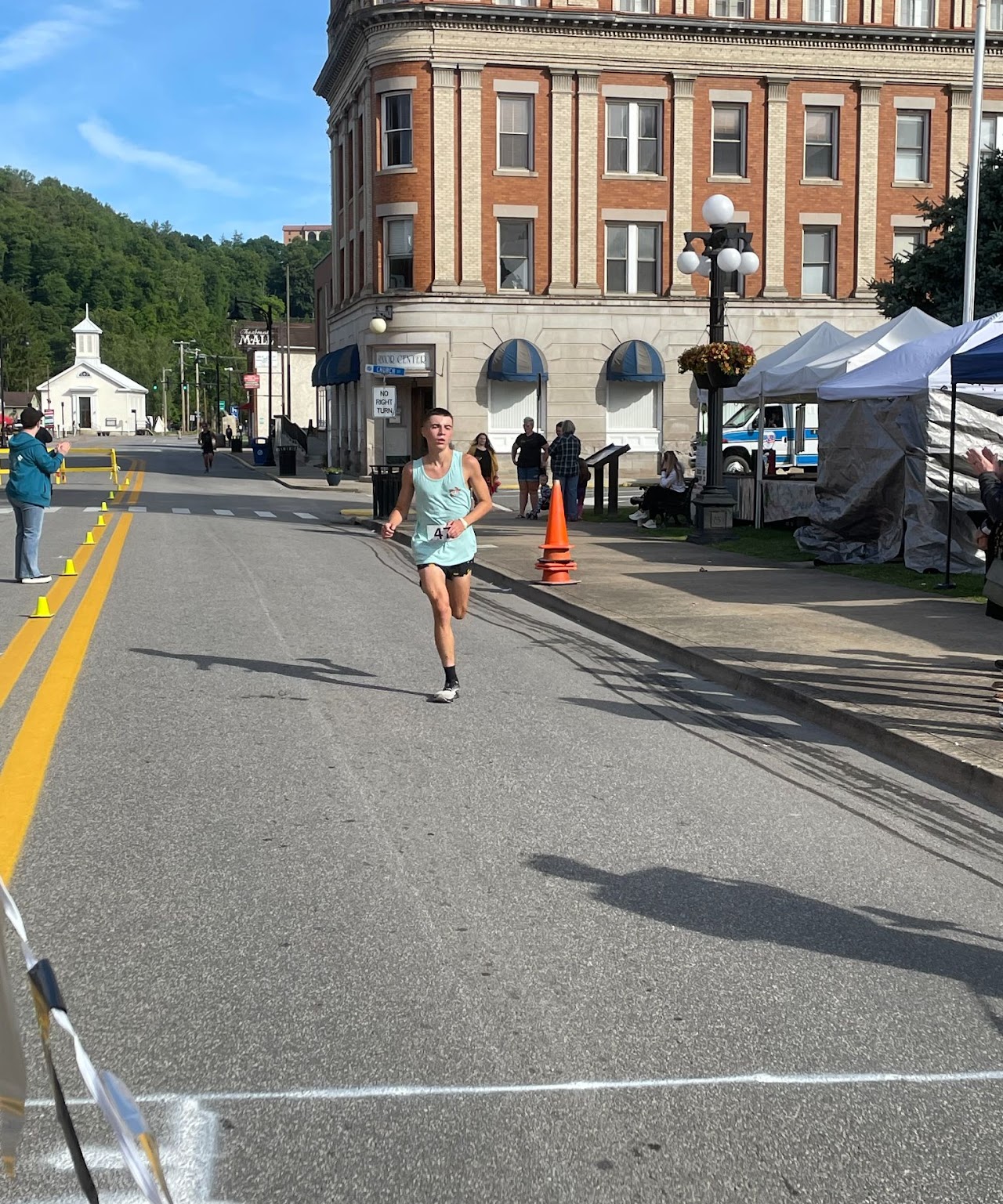 A runner in a light turquoise tank top sprints down a street lined with spectators, approaching the finish line marked by chalk.