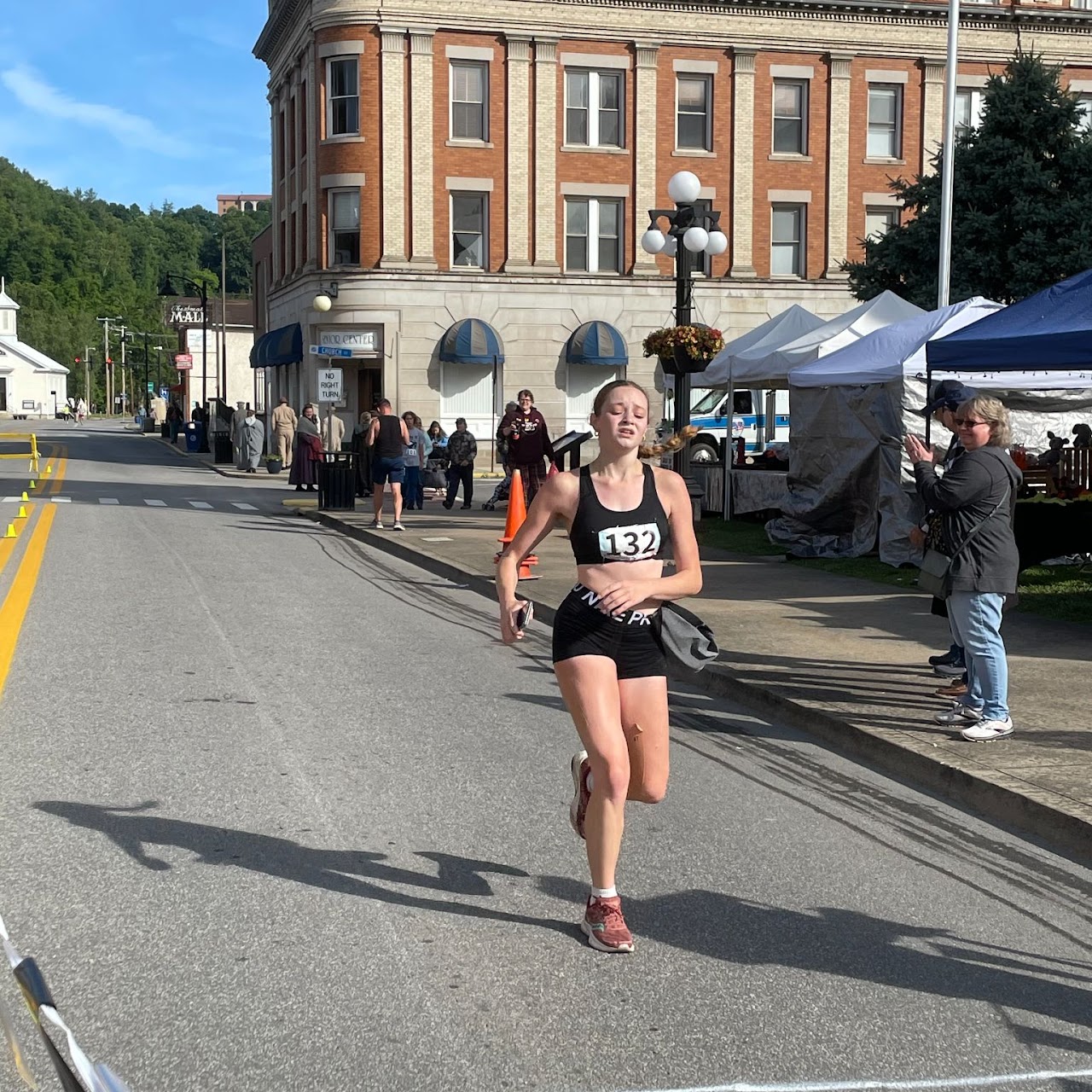 A runner in a black outfit with number 132 sprints past an audience on a street lined with tents and spectators in a sunny setting.