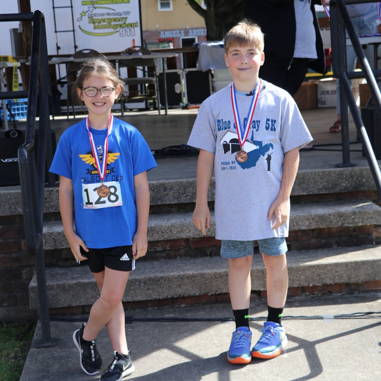 Two children stand proudly on a stage, wearing medals after a race. They are dressed in athletic gear and smiling.